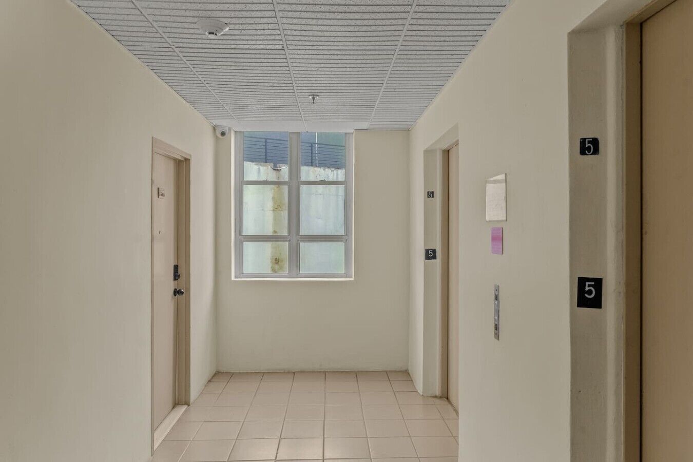 Ein Korridor mit Fliesenboden, einem großen Fenster und nummerierten Türen, darunter die Nummer 5.