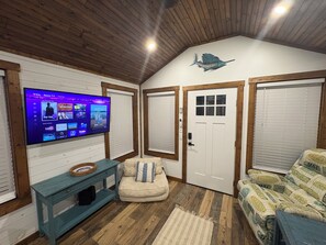 A room with a television displaying a streaming service interface, a wooden ceiling, a white door with a window, and a patterned sofa.