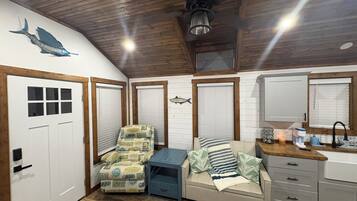 A room with a wooden ceiling, a bed with striped bedding, a chair with a beach pattern, a small table, and a window with blinds.
