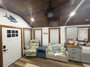 A room with a wooden ceiling, a bed with striped bedding, a chair with a beach pattern, a small table, and a window with blinds.