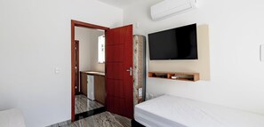 A room with a bed, a TV mounted on the wall, a small wooden shelf, and a door leading to another room. - Duplex em Porto Seguro a 10 min a Pe do Axe Moi (Porto Seguro)