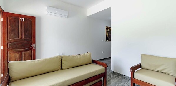 A room with a wooden door, a beige sofa, a wooden chair, and a white air conditioning unit on the wall. - Duplex em Porto Seguro a 10 min a Pe do Axe Moi (Porto Seguro)