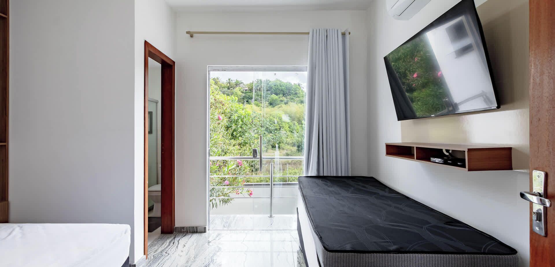 Ein Hotelzimmer mit Bett, einem an der Wand befestigten Fernseher, einem Balkon mit Blick auf Grünflächen und einer Tür zu einem weiteren Zimmer.
