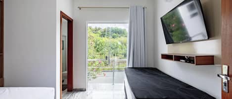Ein Hotelzimmer mit Bett, einem an der Wand befestigten Fernseher, einem Balkon mit Blick auf Grünflächen und einer Tür zu einem weiteren Zimmer.