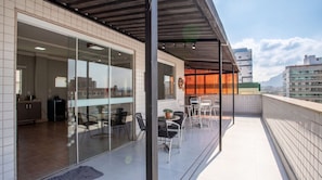 A modern building with a glass facade, a covered outdoor seating area with tables and chairs, and a view of the cityscape. - Cobertura a 40m da Praia do Morro c Churrasqueira (Luis Correia)