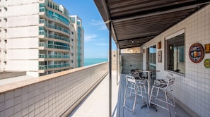 A balcony with a view of the ocean, a bar area with stools, and a building with multiple balconies. - Cobertura a 40m da Praia do Morro c Churrasqueira (Luis Correia)
