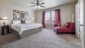 Quarto com cama, ventilador de teto, janela com cortinas, cadeira e mesa de cabeceira com abajur.