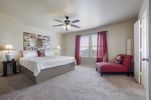 Une chambre à coucher comprenant un lit, un ventilateur de plafond, une fenêtre avec des rideaux, une chaise et une table de chevet avec une lampe.