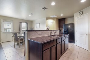 A kitchen with a dark brown countertop, a sink, and a refrigerator. There is a dining table with chairs and a window with blinds. - Family Time - 12 min. to Downtown & The Riverwalk (San Antonio)