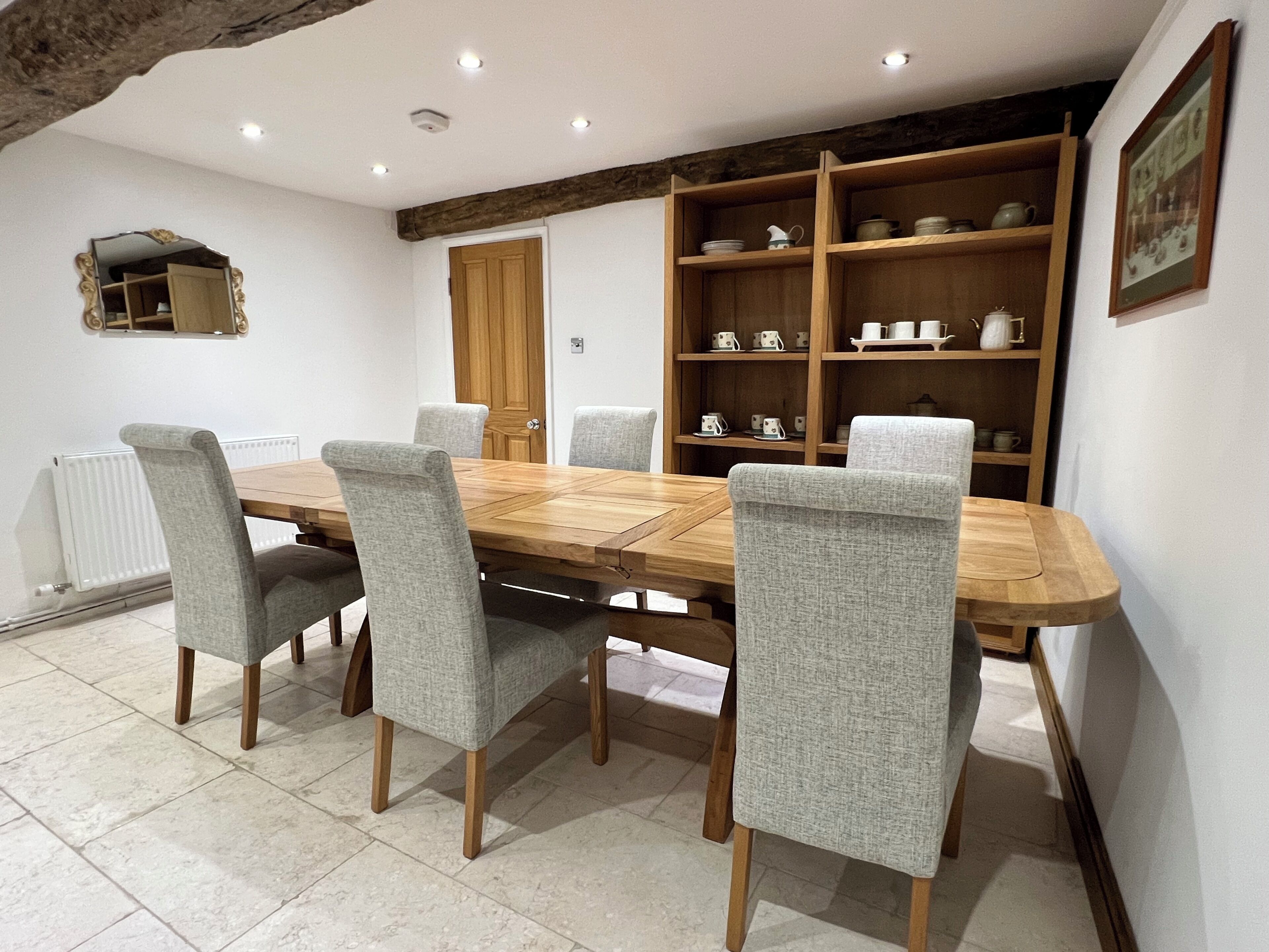 A dining area with a wooden table and grey upholstered chairs, a wooden bookshelf with tea sets, and a white radiator.
