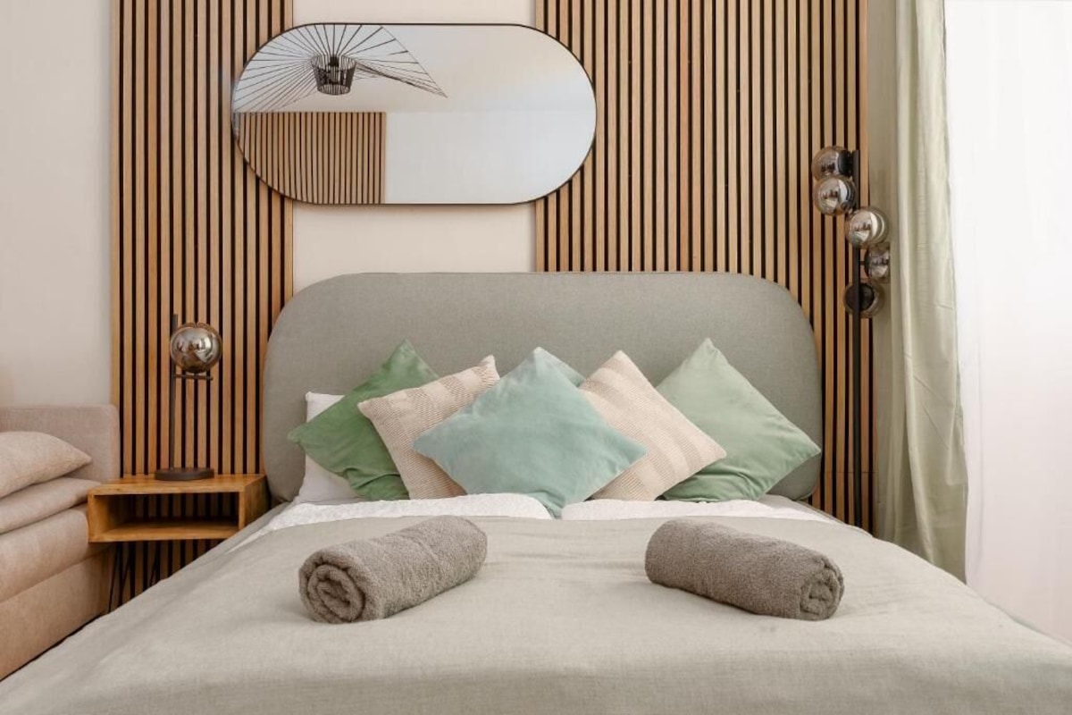 A neatly made bed with rolled towels, a bedside table with a lamp, and a wall-mounted mirror.