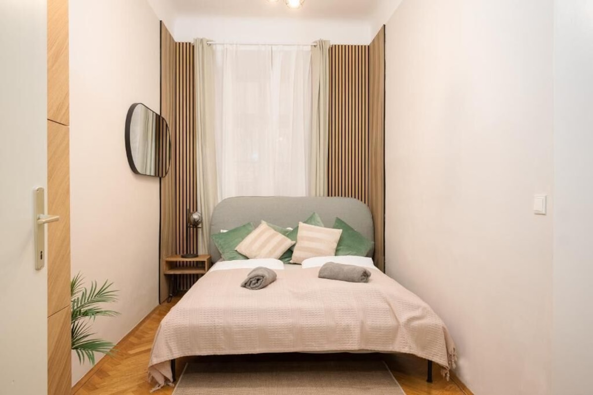 A narrow hotel room with a bed, a bedside table, a mirror, and a window with curtains.