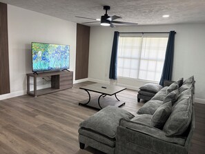Ein modernes Wohnzimmer mit Fernseher, Deckenventilator, Esstisch und grauem Sofa.