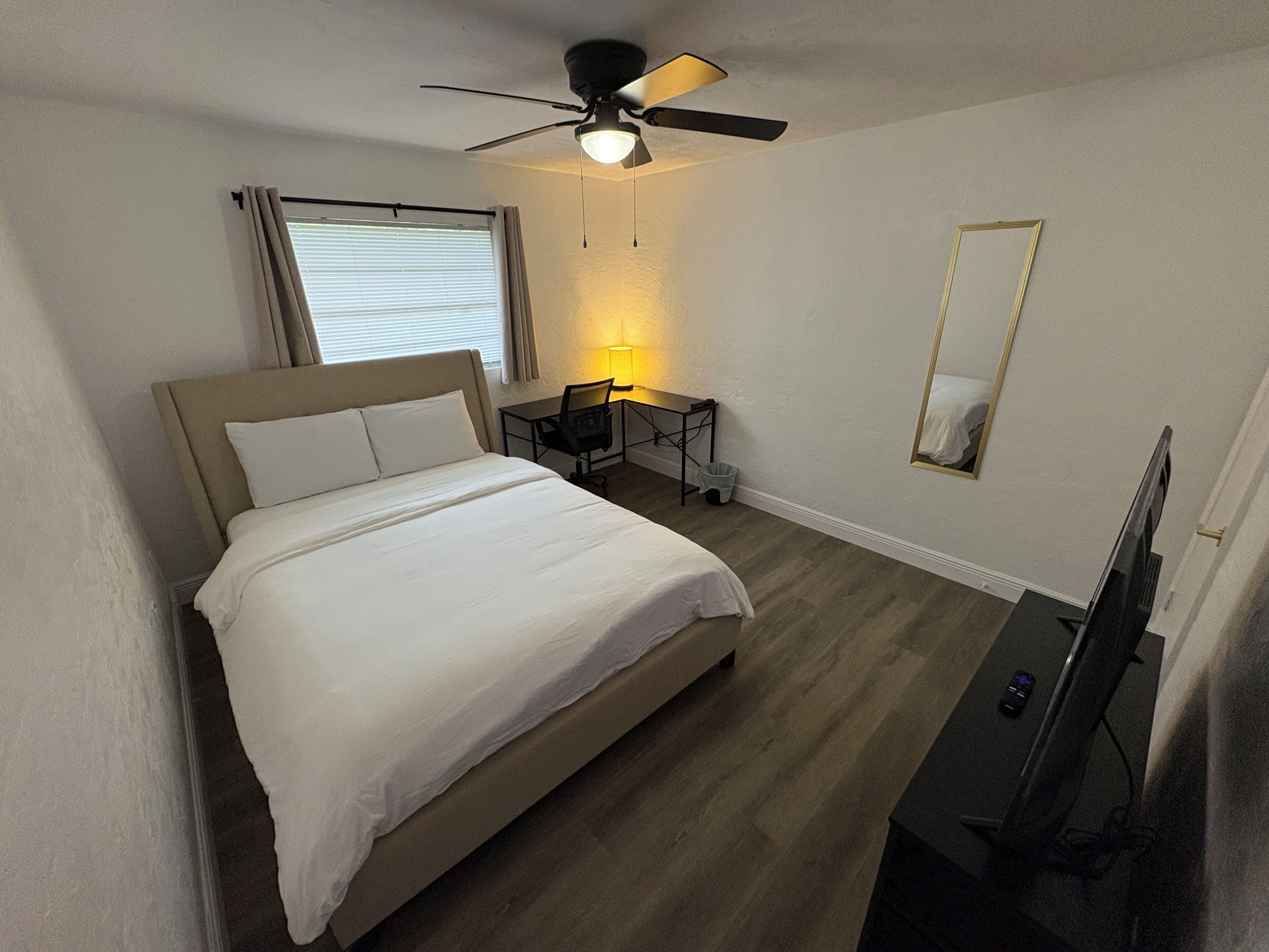 A hotel room with a bed, a desk, a chair, a television, a ceiling fan, and a window with curtains.