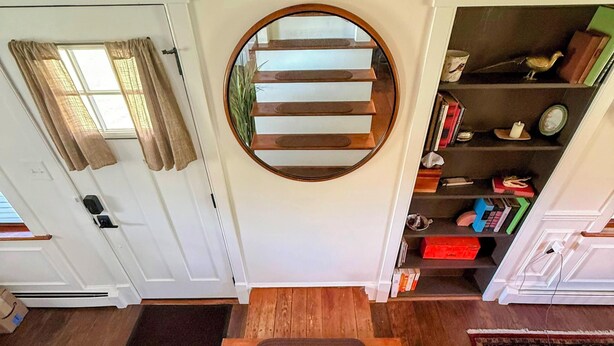 A staircase with a circular mirror above it, leading to a landing with a bookshelf and a door with curtains.