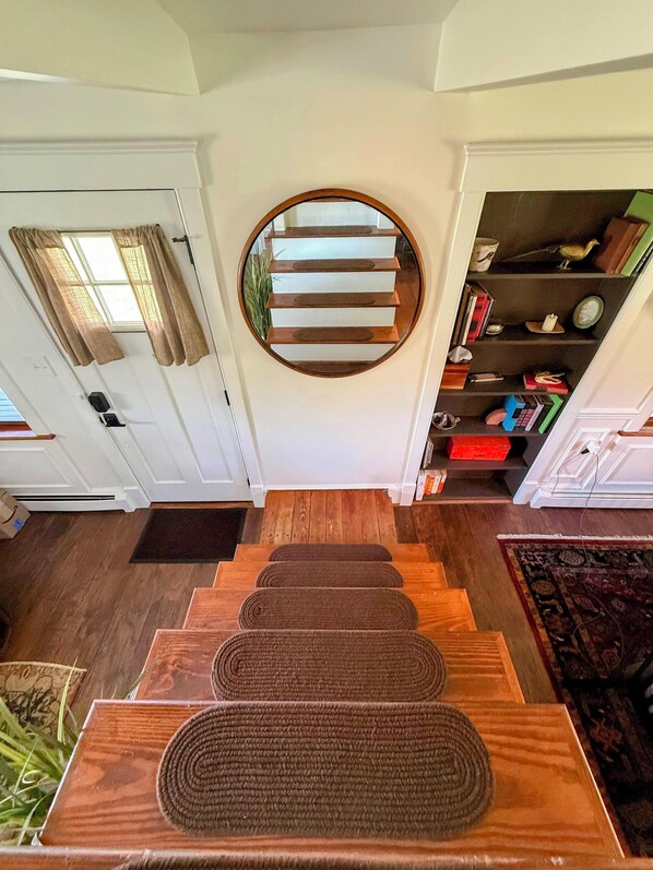 A staircase with a circular mirror above it, leading to a landing with a bookshelf and a door with curtains.