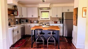 A modern kitchen with white cabinets, stainless steel appliances, a wooden island with bar stools, and a window with blinds.