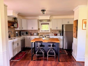 A modern kitchen with white cabinets, stainless steel appliances, a wooden island with bar stools, and a window with blinds.
