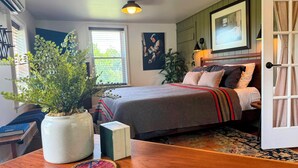 Quarto com cama, mesa de cabeceira, planta em vaso, janela e um quadro na parede.