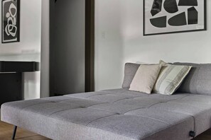 Ein graues Schlafsofa mit einer gesteppten Optik, zwei Kissen und ein schwarzer Metallhocker.