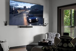 Ein Wohnzimmer mit einem großen Flachbildfernseher an der Wand, einem Sofa, einem Couchtisch mit Würfeln und einem Fenster mit Blick auf Grünflächen.