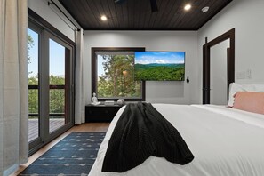 Un dormitorio con una cama grande, un televisor montado en la pared, un ventanal con vistas a la vegetación y una mesita de noche pequeña.