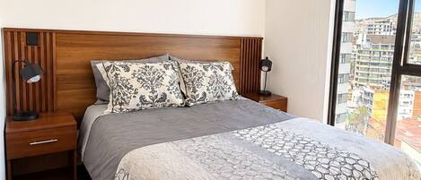 A bedroom with a wooden bed, a nightstand, a lamp, and a large window with a city view.