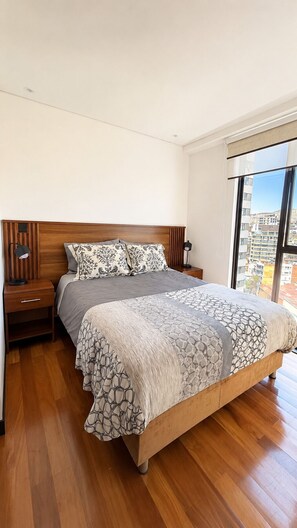 Ein Schlafzimmer mit einem hölzernen Bett, einem Nachttisch, einer Lampe und einem großen Fenster mit Blick auf die Stadt.