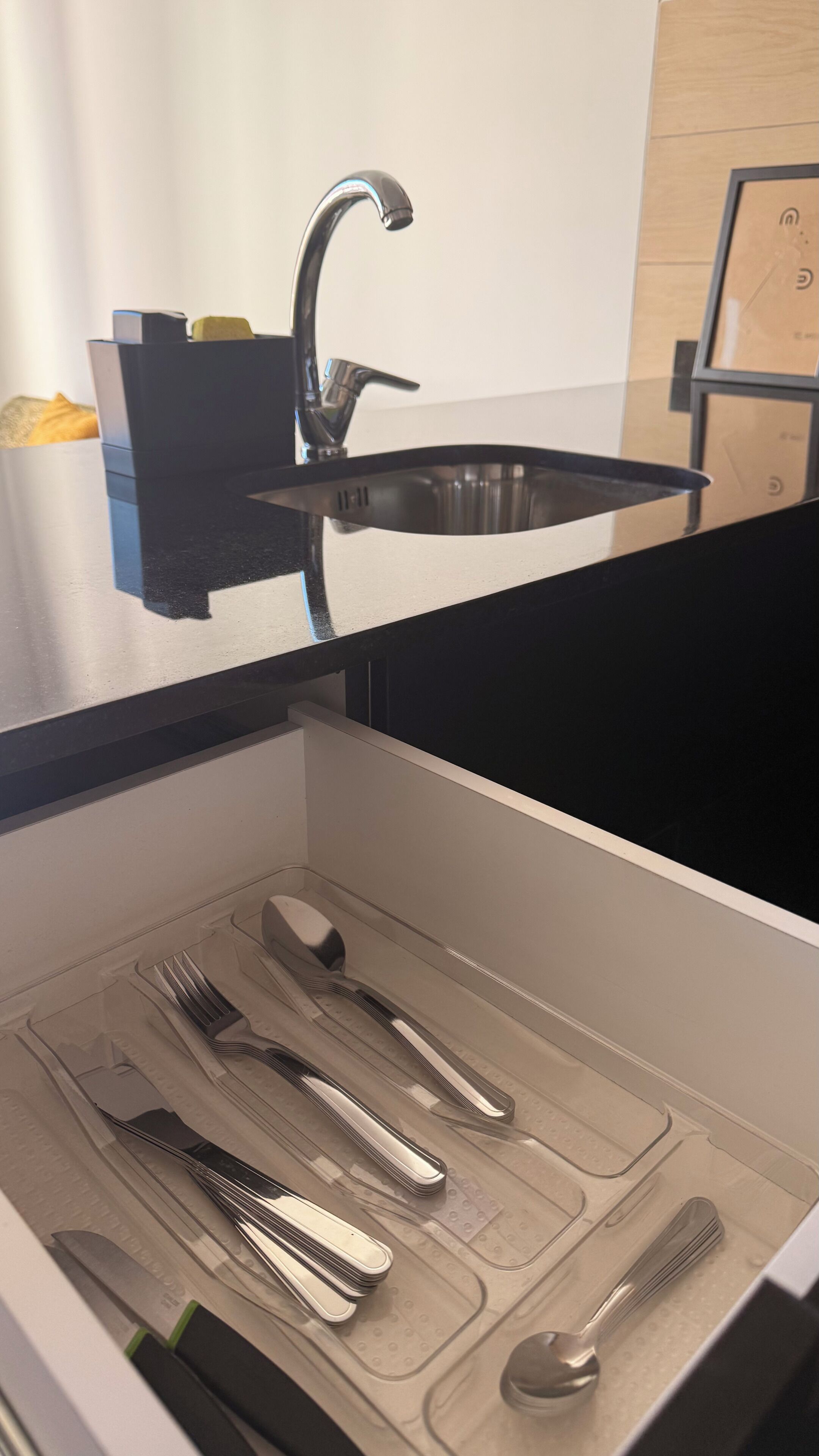 Une cuisine dotée d’un évier en acier inoxydable, d’un plan de travail noir et d’un tiroir contenant des ustensiles rangés avec soin.