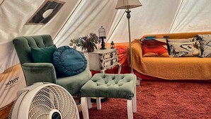 Ein Zeltinnenraum mit einem grünen Sessel, einem kleinen Hocker, einem Standventilator, einer Lampe und einer Couch mit gemusterten Kissen.