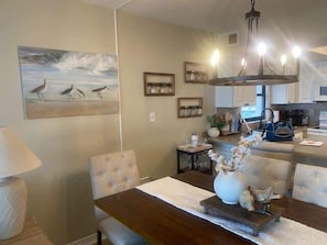 A dining area with a wooden table, chairs, and a vase with flowers. There is a painting on the wall and a chandelier above the table. - Sunswept 508 by ALBVR (Orange Beach)