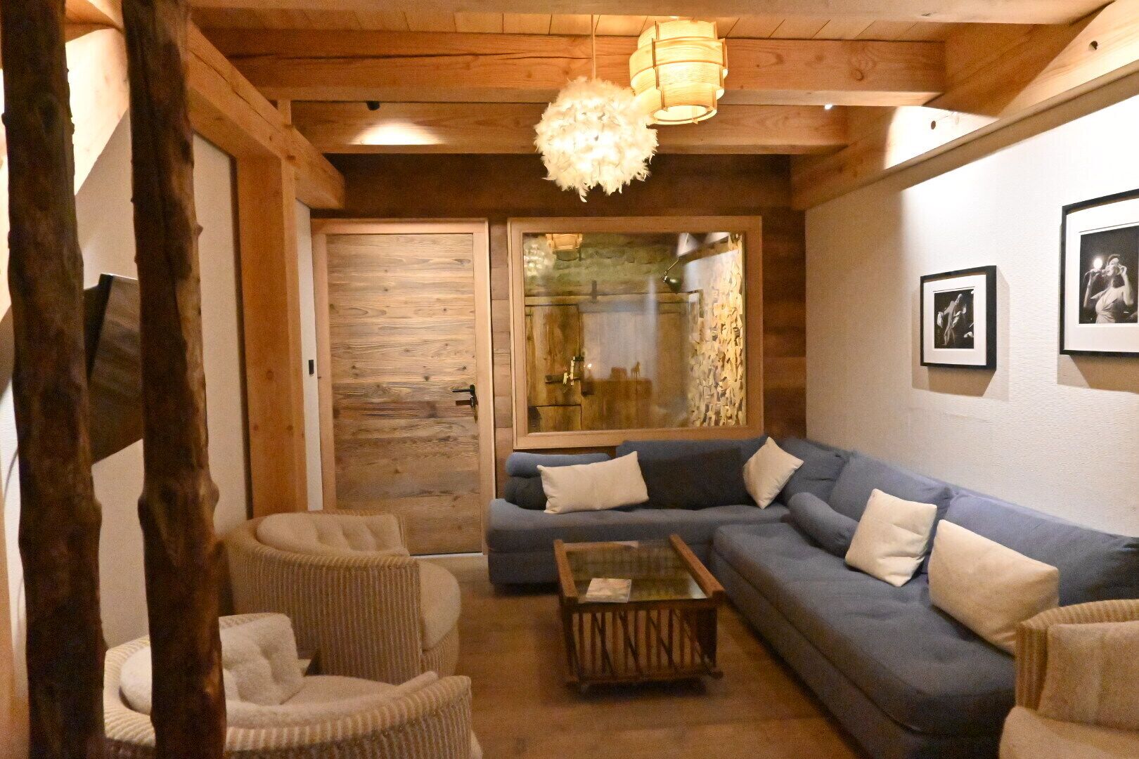 A cozy living room with a blue sectional sofa, two armchairs, a coffee table, wooden beams, a wooden door, and a large window with a view.