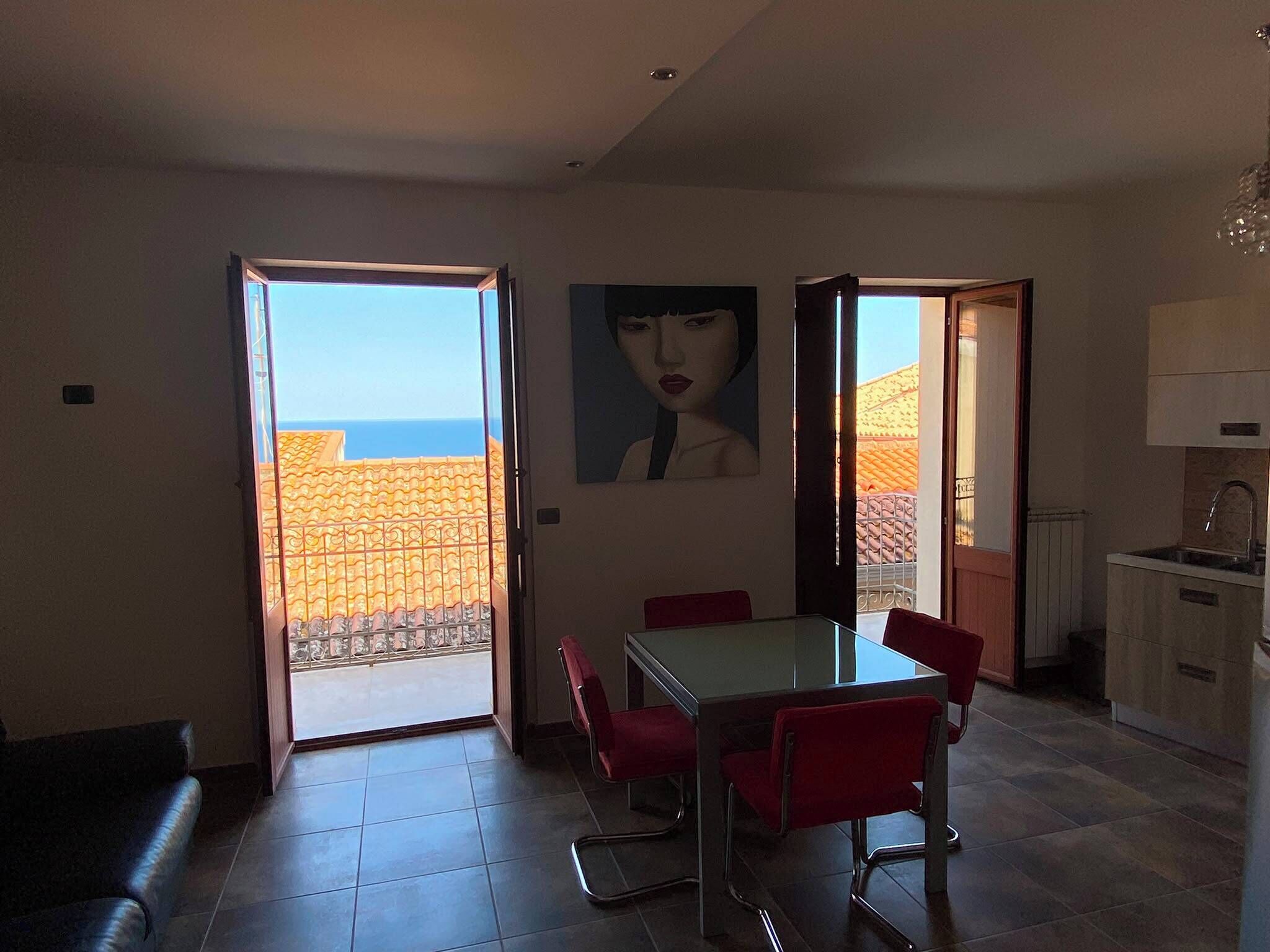 Un coin repas avec une table et des chaises, un tableau accroché au mur et une vue sur la mer par des portes ouvertes.