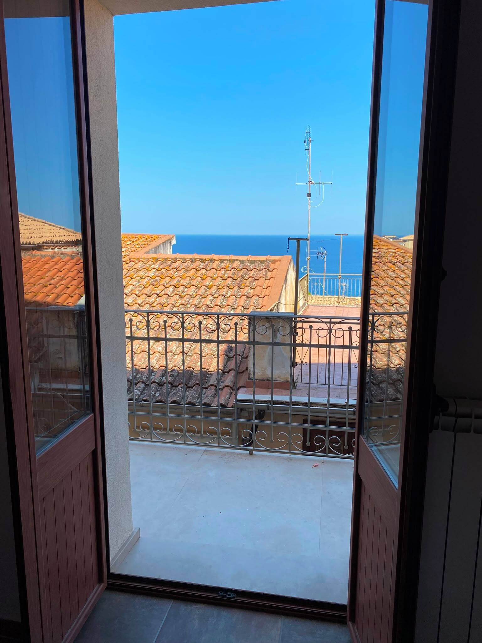 Un balcone con vista su una zona costiera, con tetti in terracotta e un cielo azzurro.