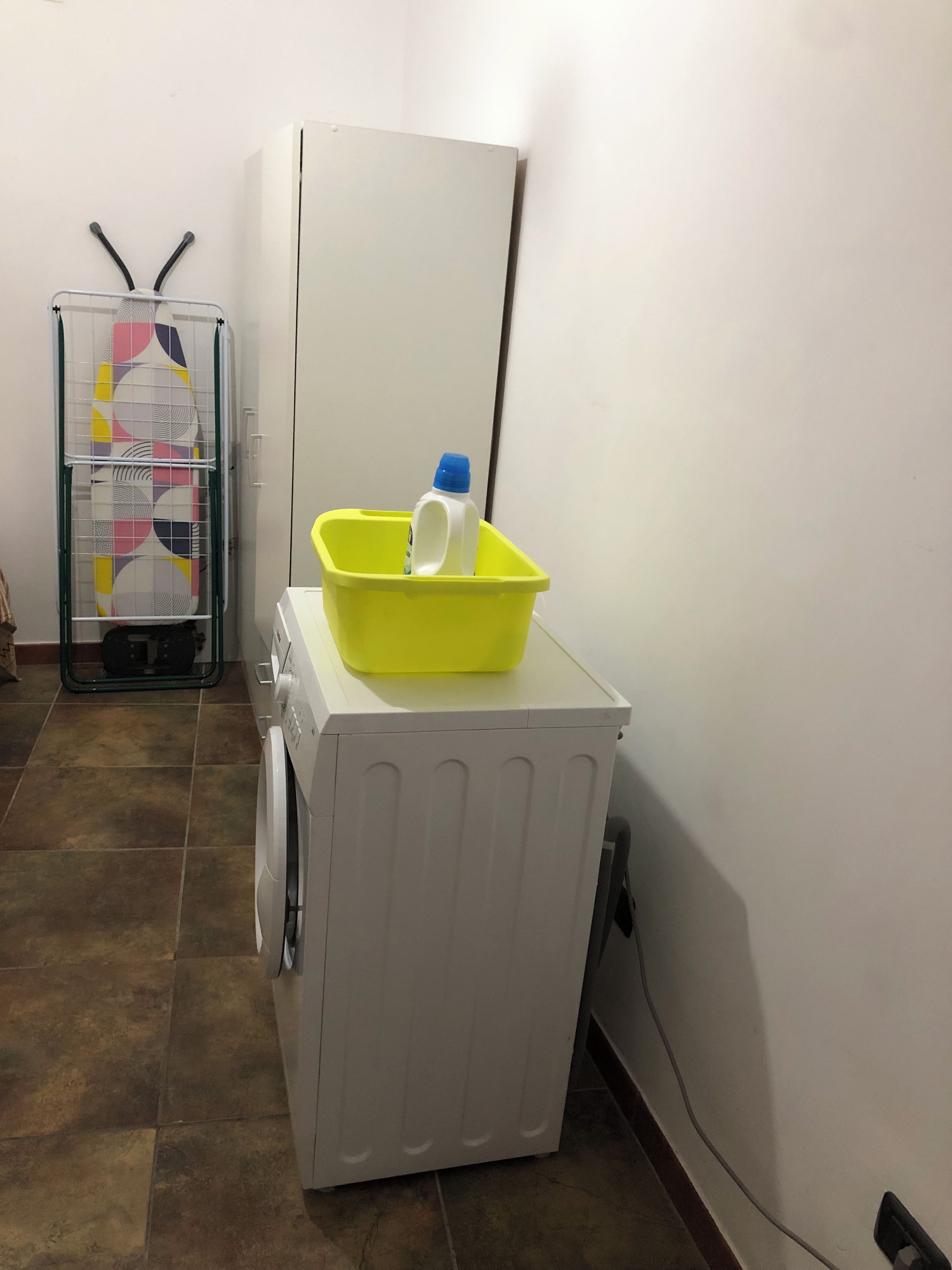 Ein Waschbereich mit Waschmaschine, einem gelben Becken darauf und einem Wäscheständer mit gemustertem Stoff.