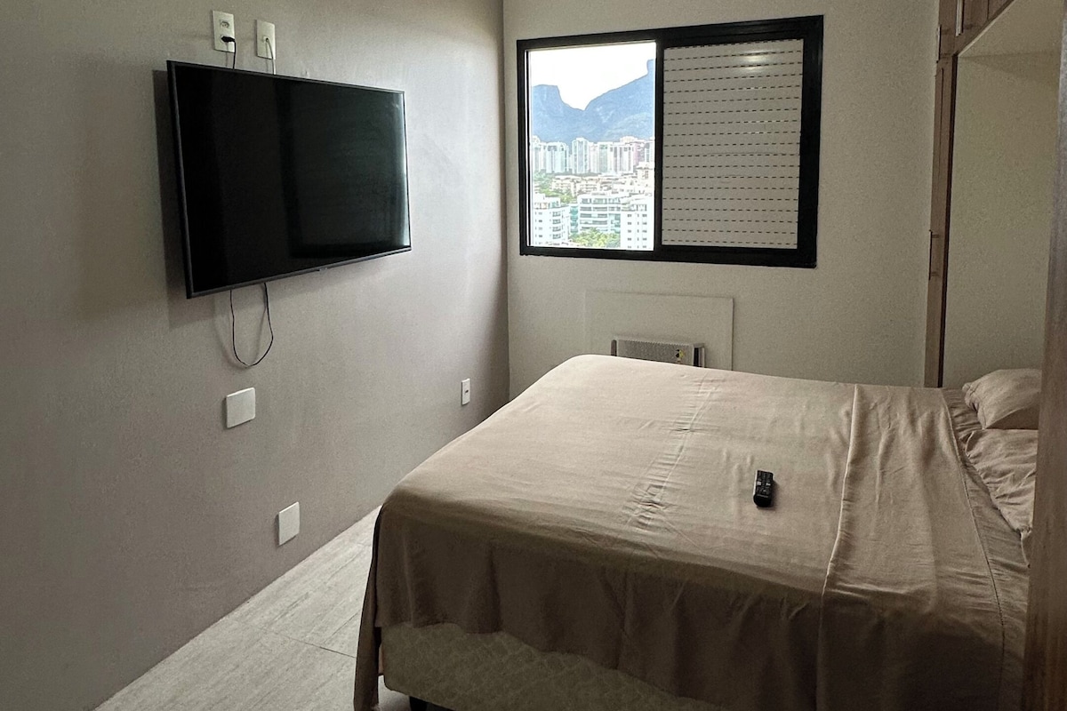 Een slaapkamer met een bed, een plafondventilator, een aan de muur bevestigde televisie en een raam met uitzicht op gebouwen.