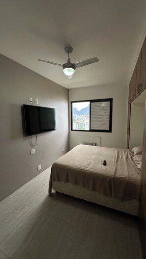 Een slaapkamer met een bed, een plafondventilator, een aan de muur bevestigde televisie en een raam met uitzicht op gebouwen.