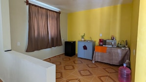 A room with a sloped ceiling, a window with brown curtains, a kitchenette with a table, and a water dispenser.