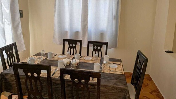 A dining area with a table set for four, surrounded by chairs, with a window and curtains in the background.