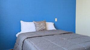 A bed with a grey bedspread and two pillows, one plain and one with a wavy pattern, against a blue wall with a tiled floor.