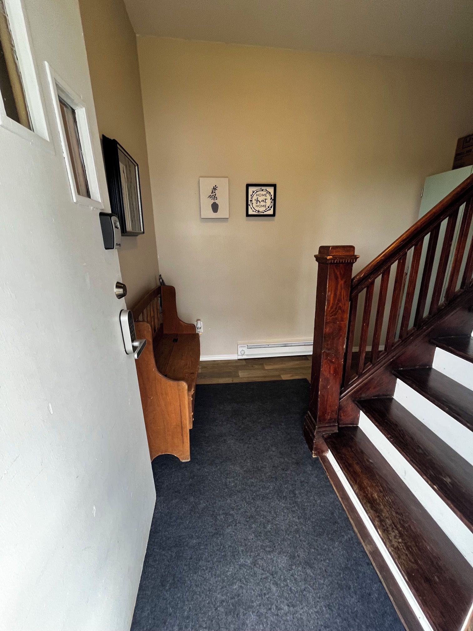 Ein Flur mit einer Holztreppe, einer Tür mit Griff, einer kleinen Holzbank und zwei gerahmten Bildern an der Wand.