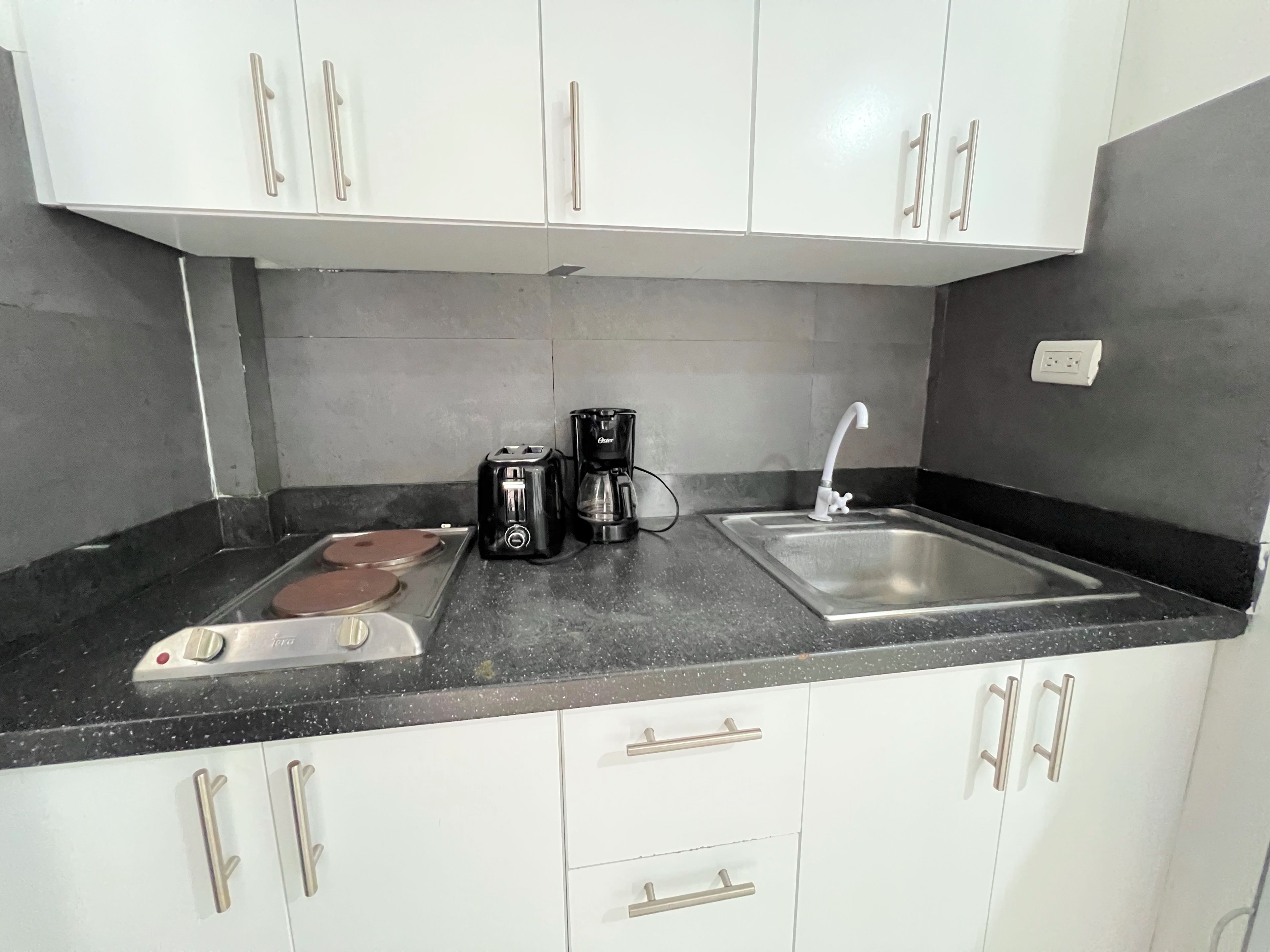 Una cocina con armarios blancos, encimera negra, cafetera, tostadora y un lavamanos.
