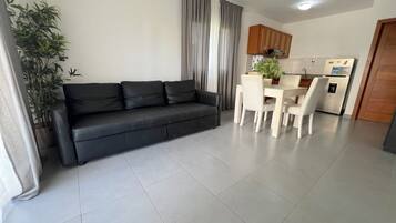 Una sala moderna con un sofá negro, una mesa de comedor con sillas, una zona de cocina con armarios y un refrigerador, y una planta en maceta.