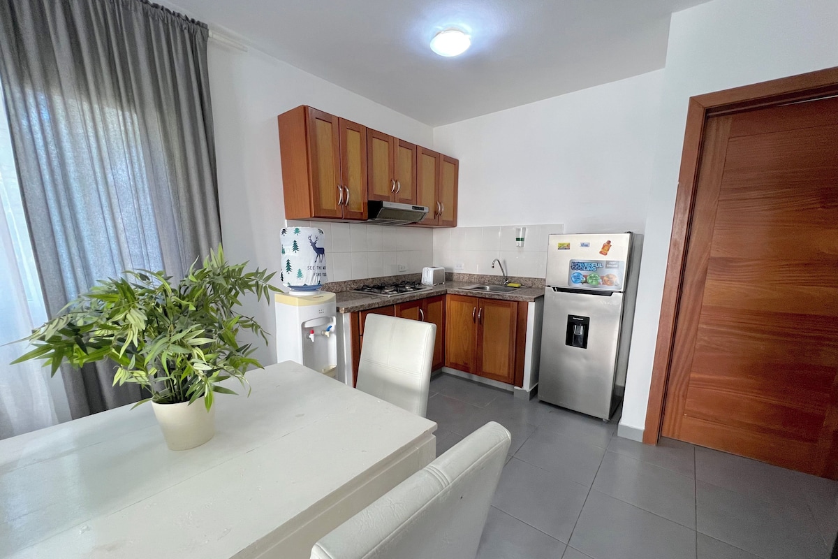 A compact kitchen with wooden cabinets, a refrigerator, a water dispenser, and a dining table with chairs.