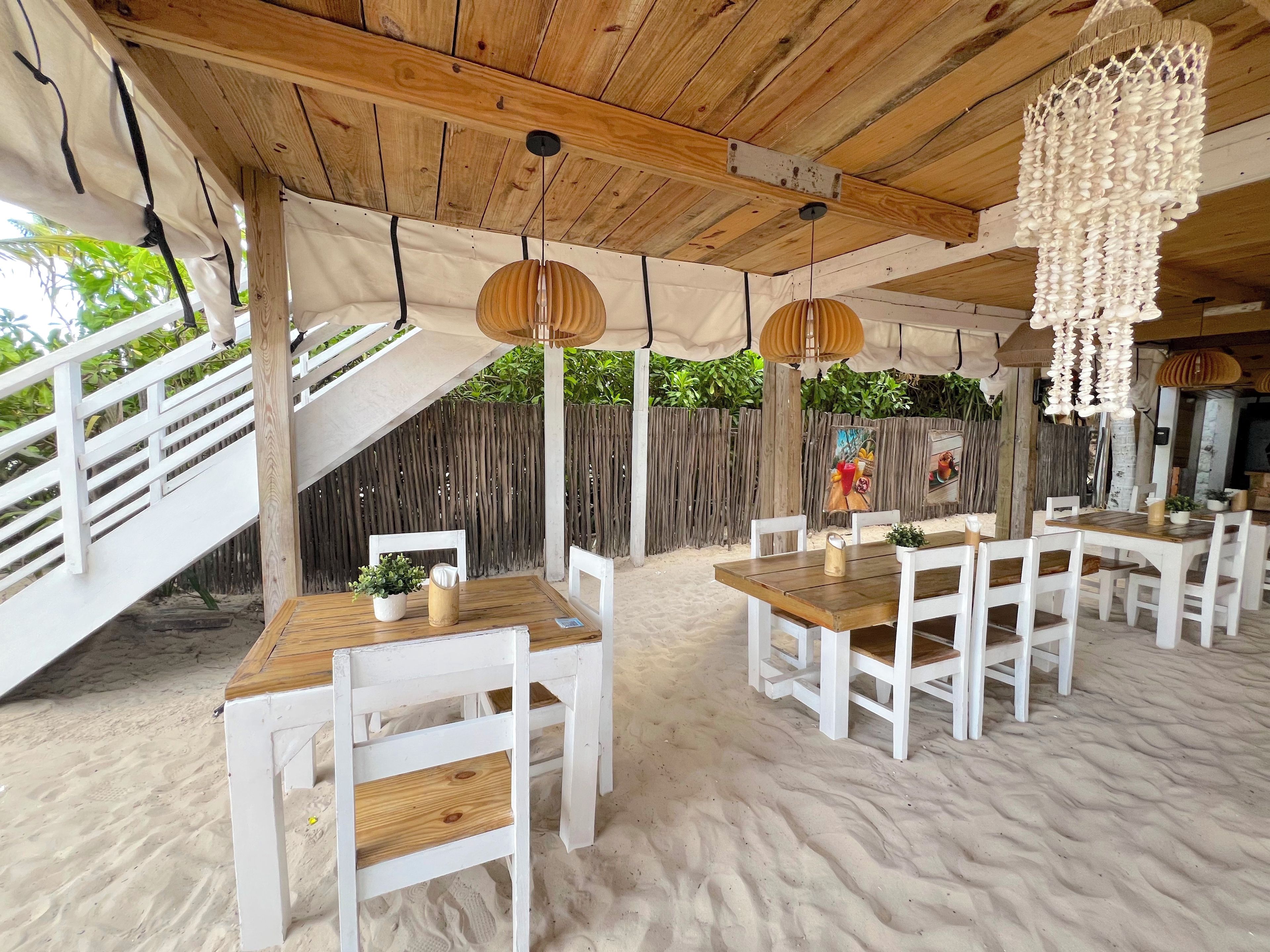 Un espacio para comer al aire libre con sillas blancas y mesas de madera, techo de madera y suelo de arena.
