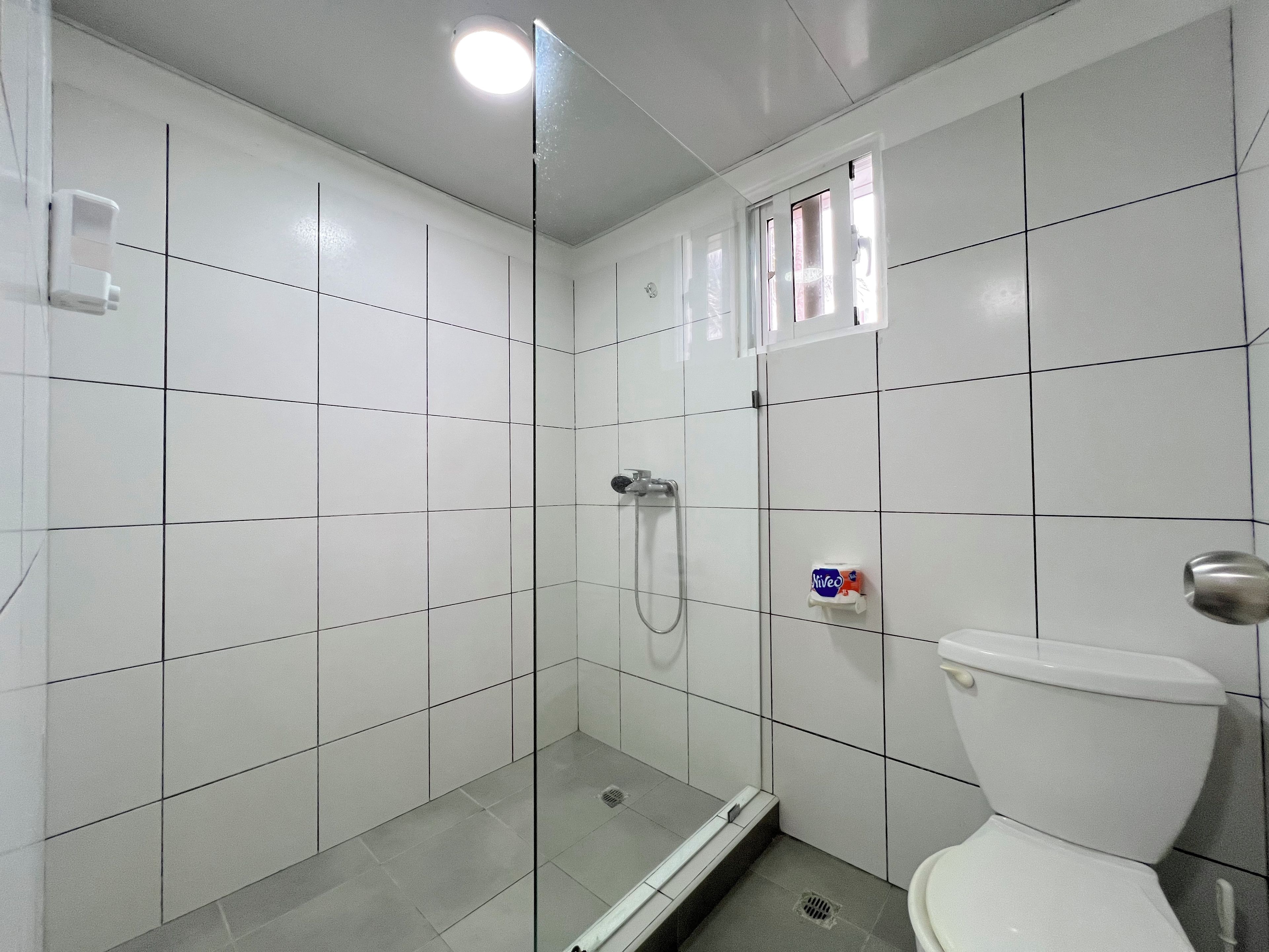 Ein Badezimmer mit weißen Fliesen, einer Glasduschkabine, einer Toilette und einem kleinen Fenster.