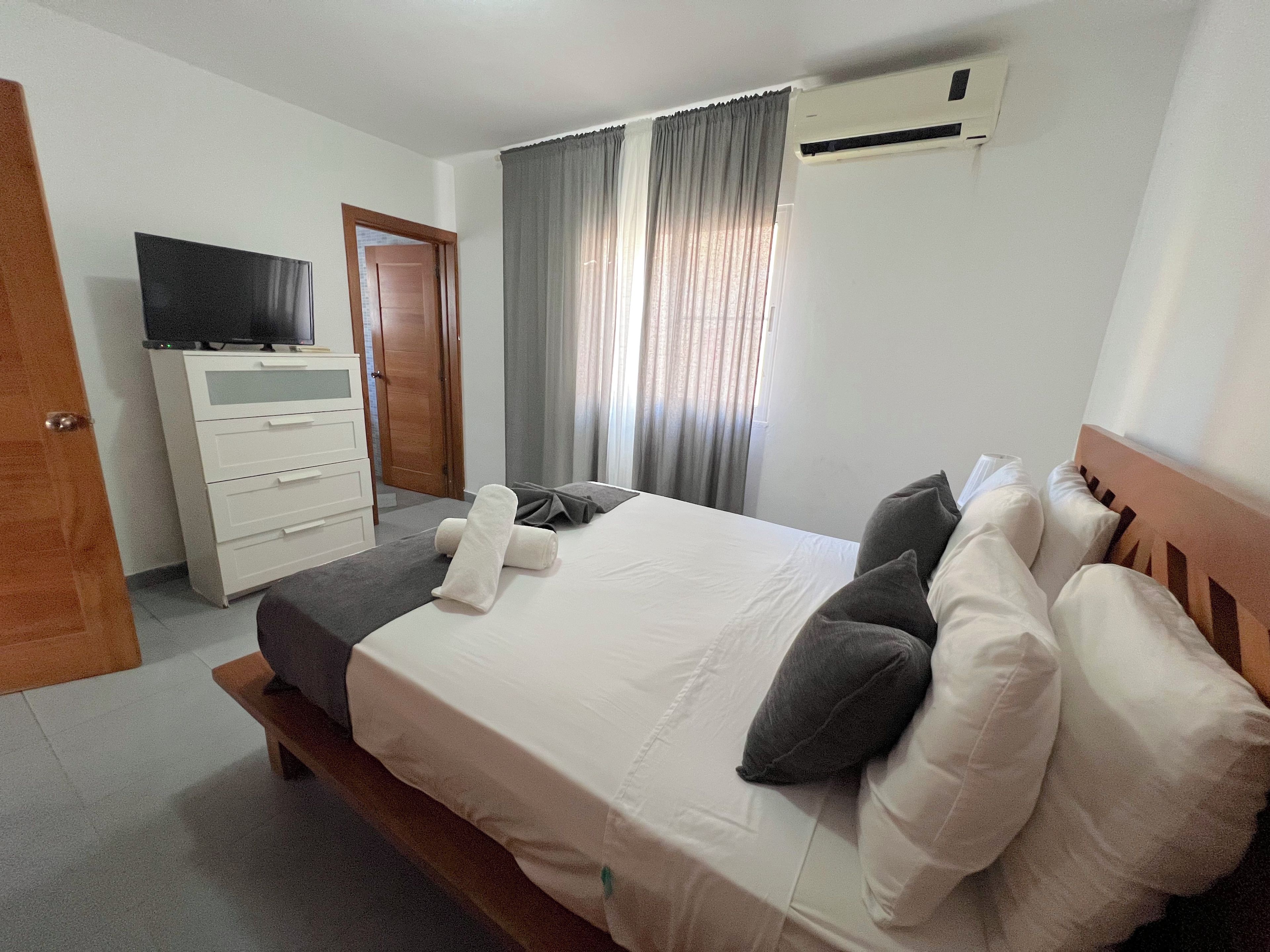 A hotel room with a bed, a TV on a dresser, a door, and an air conditioning unit.