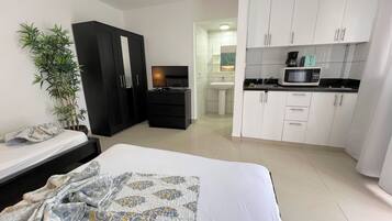 Ein Schlafzimmer mit Bett, Fernseher, Kleiderschrank, Schreibtisch, Spüle und einer Mikrowelle in der angrenzenden Küchenzeile.