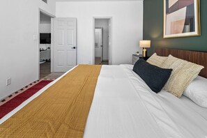 Una habitación de hotel con una cama, una mesita de noche con lámpara, una alfombra roja y blanca, y una puerta que da a un pasillo.