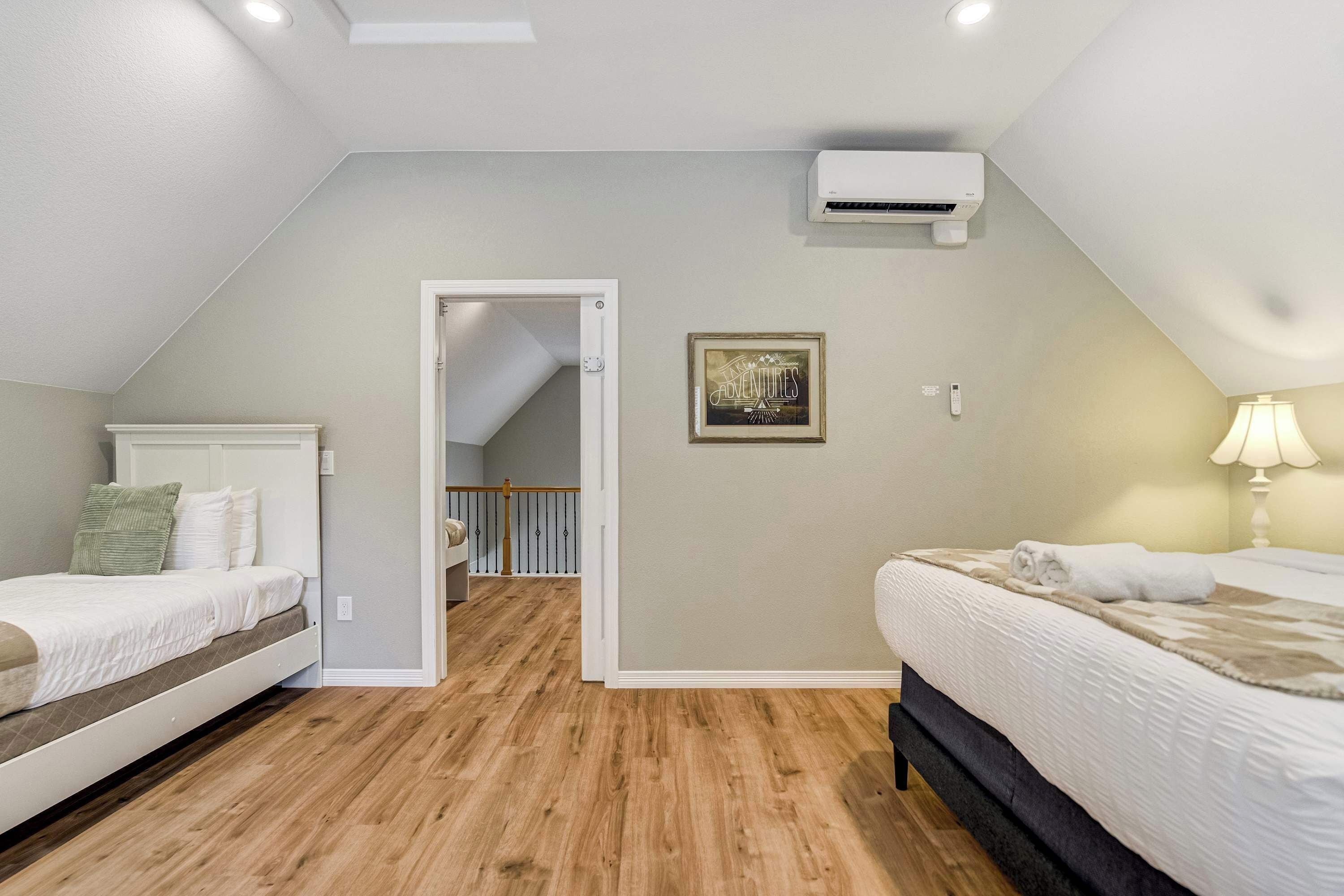 A bedroom with two beds, a ceiling fan, an air conditioning unit, a framed picture, and a door leading to a staircase.